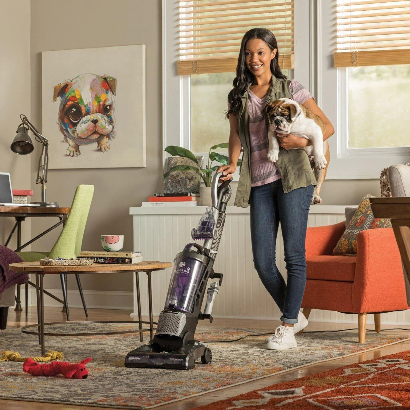 A woman vacuums using her purple and silver vacuum while holding her bulldog in her living room